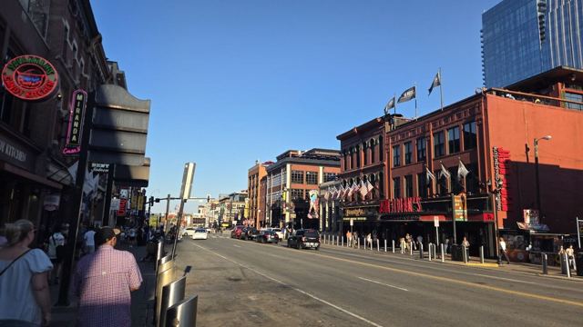 A sunny street of colourful bars and honky-tonks in downtown Nashville with tall buildings behind.