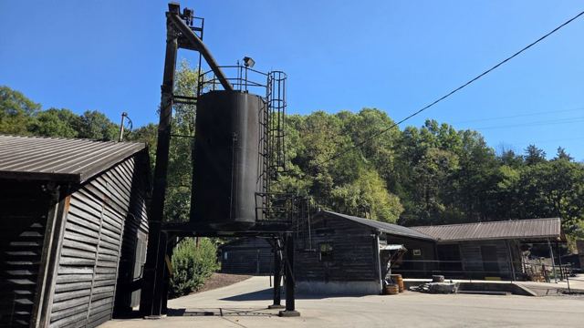 Black wooden distillery buildings and a water tower among green trees under a blue sky.