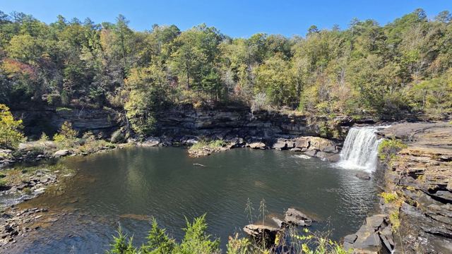 A small waterfall pouring into a wide pool surrounded by rocky cliffs and forest.