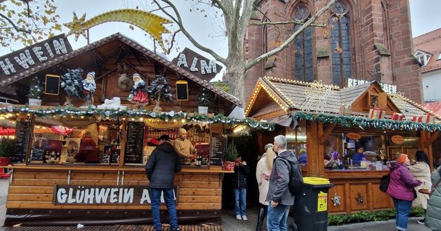 A wooden Glühwein stall decorated for Christmas, with visitors and a red church behind.