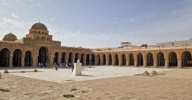 Large paved mosque courtyard surrounded by arched arcades with a dome in the background.
