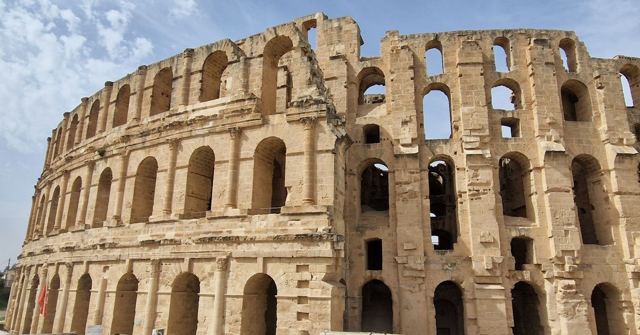 Exterior of a huge stone Roman amphitheatre with tiers of arches against a blue sky.