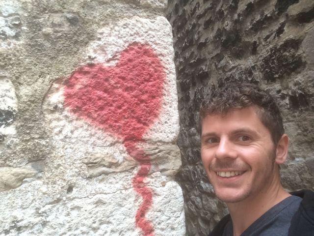 A smiling man taking a selfie beside a red heart painted on a stone wall in Split.