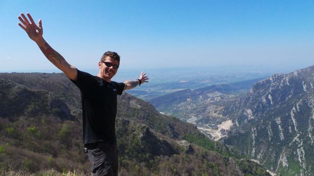 A man waving with arms out at a mountain lookout above a hazy valley in Albania.