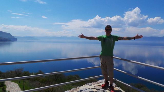 A man with arms outstretched by a railing above calm blue Lake Ohrid.