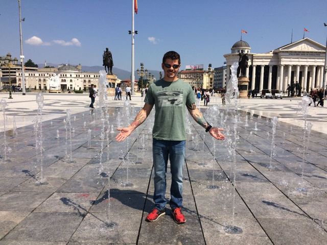A man with arms out standing among water fountains in Macedonia Square in Skopje.