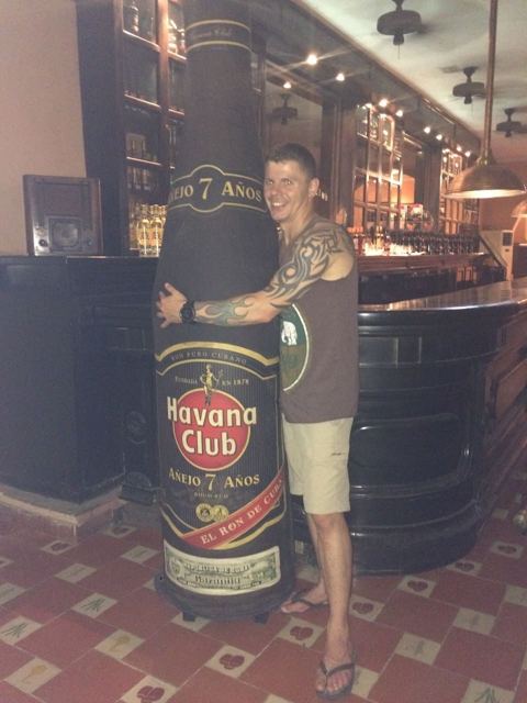 A man posing next to a giant Havana Club rum bottle in a bar.
