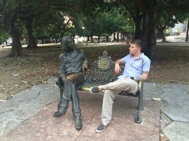 A man sitting on a park bench beside a bronze statue of John Lennon.