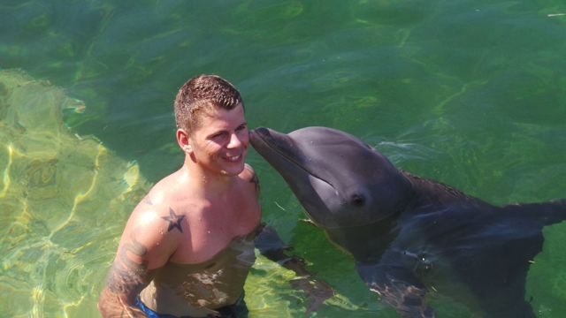 A man in the water being kissed on the cheek by a dolphin.