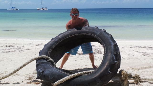 A man lifting a large tractor tyre on a white sand beach by turquoise sea.