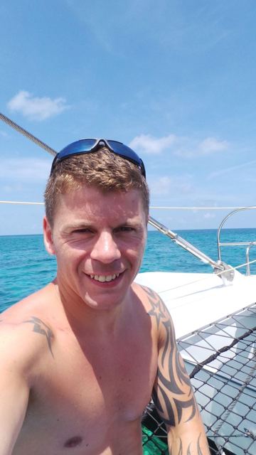 A smiling man with sunglasses on his head on a catamaran deck above turquoise water.