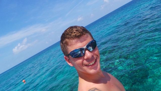 A smiling man in sunglasses taking a selfie above clear turquoise sea.