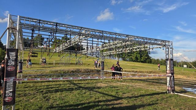 Large metal obstacle rig on a grassy slope with people nearby on a sunny day.