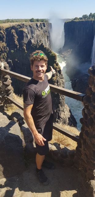 A smiling man leaning on a railing with the gorge and waterfall behind.
