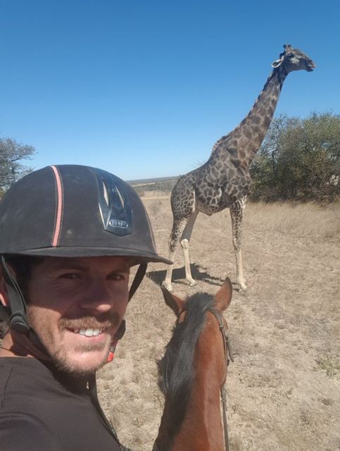 A man in a riding helmet taking a selfie on horseback with a giraffe behind.