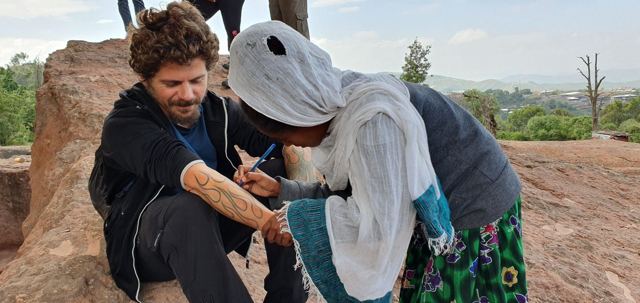 A man sitting on rocks while a woman in a white shawl draws on his arm.