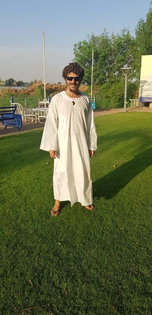 A man in sunglasses wearing a long white jalabiya on a lawn.