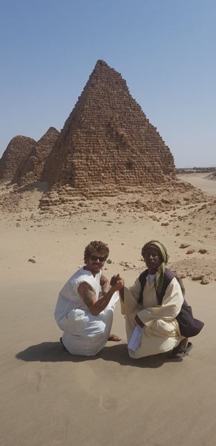 A man in a white top kneeling beside a local man in front of ancient Nubian pyramids in the desert.