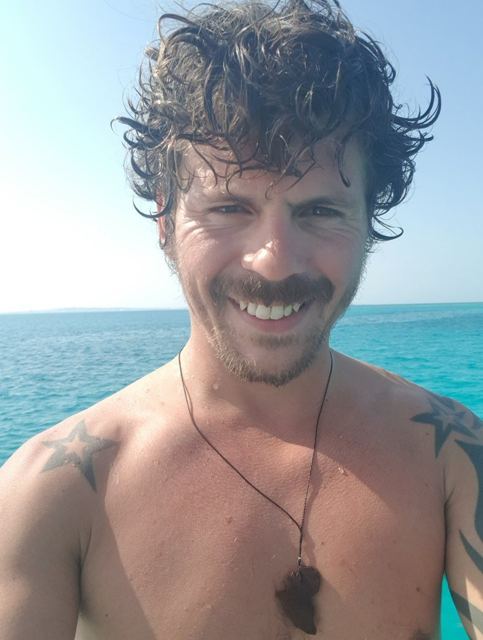 A smiling shirtless man with wild curly hair taking a selfie in front of turquoise water.