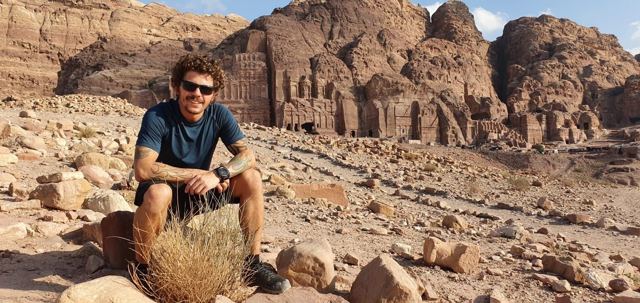 A man in sunglasses sitting on rocks in front of the carved Royal Tombs at Petra.