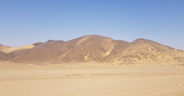 Barren brown hills rising from a flat sandy desert under a clear blue sky.