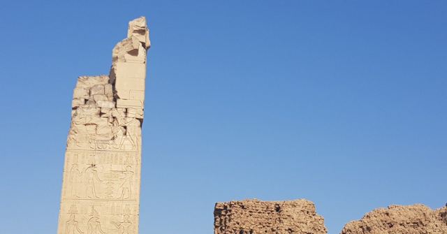 A single weathered stone column standing against a bright blue sky beside low ruined walls.