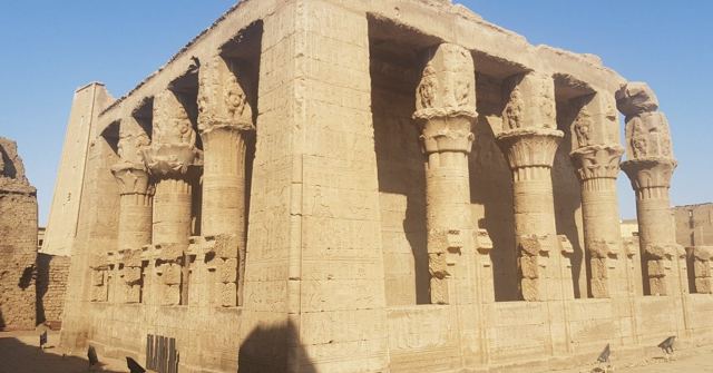 Sandstone temple facade with ornate carved column capitals and screen walls under blue sky.