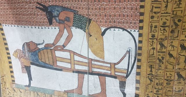 Ancient tomb painting of jackal-headed Anubis tending a mummy on a bier, surrounded by hieroglyphs.