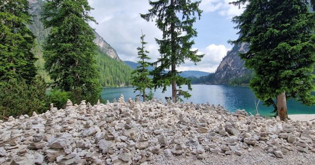 Hundreds of small stone cairns on a rocky shore with pine trees and a turquoise lake behind.