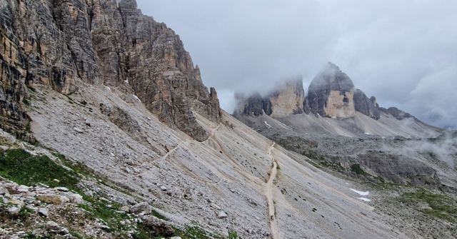 A gravel trail through a scree valley towards three rocky peaks partly covered in cloud.