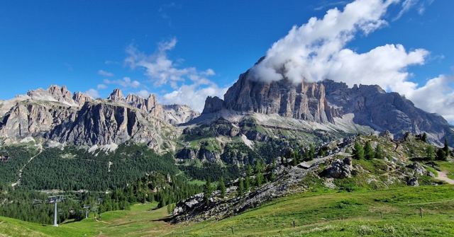 Rugged grey mountains under a blue sky with a cloud draped over one peak and green slopes below.