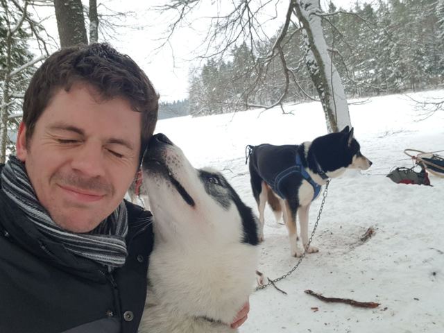 A man smiling as a husky licks his face in a snowy forest.