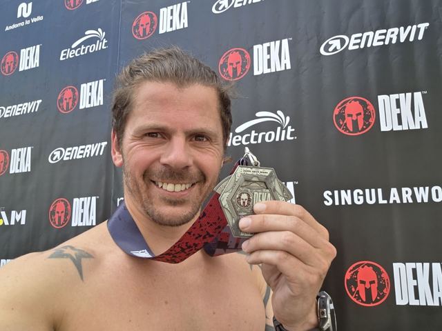 Smiling shirtless man holding up a race medal in front of a Spartan DEKA sponsor wall.