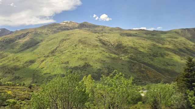 Lush green mountain slopes and trees under a blue sky with small clouds.