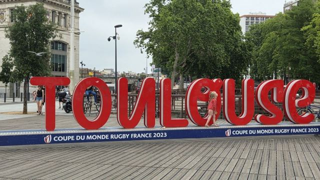 Large red letters spelling Toulouse on a wooden deck with trees and buildings behind.