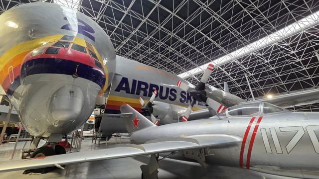 Nose of an oversized cargo plane in a hangar with a silver fighter jet beneath it.