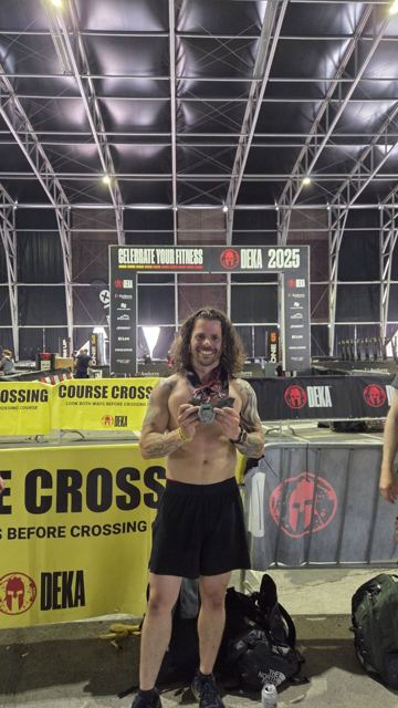 A shirtless man holding a medal under the DEKA finish arch.
