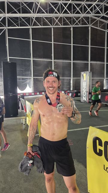 A shirtless man with a race headband showing a medal in an indoor race arena.