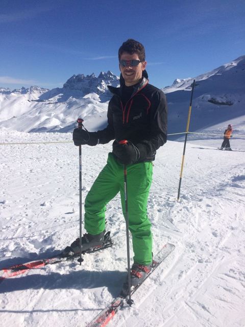 A man in green ski trousers standing on skis with poles on a sunny snowy slope.