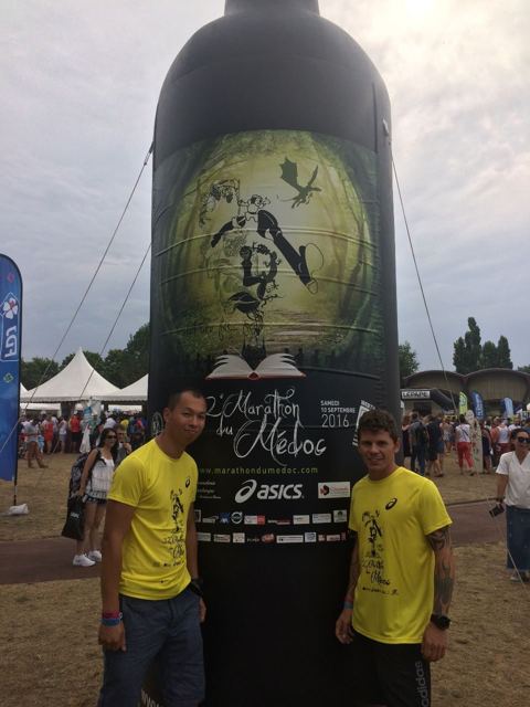 Two runners in yellow T-shirts standing in front of a giant inflatable wine bottle at the Marathon du Médoc start.