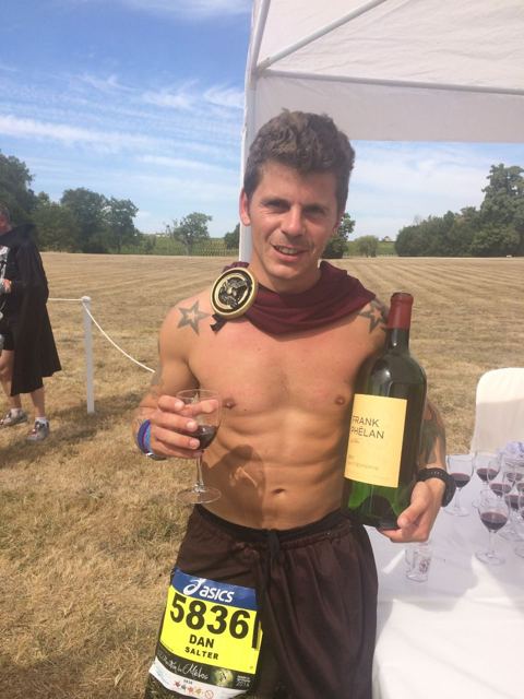A shirtless runner in a red cape with a race bib holding a glass and a bottle of wine in a vineyard.