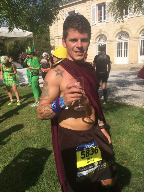 A runner in a red cape and race bib holding a glass of wine on the lawn of a château.