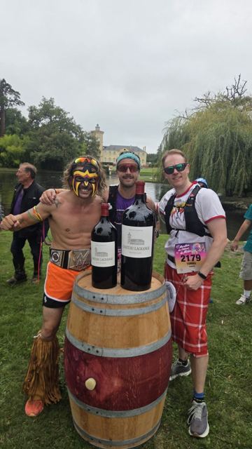 Runners in costume posing with wine bottles on a barrel in a château garden.