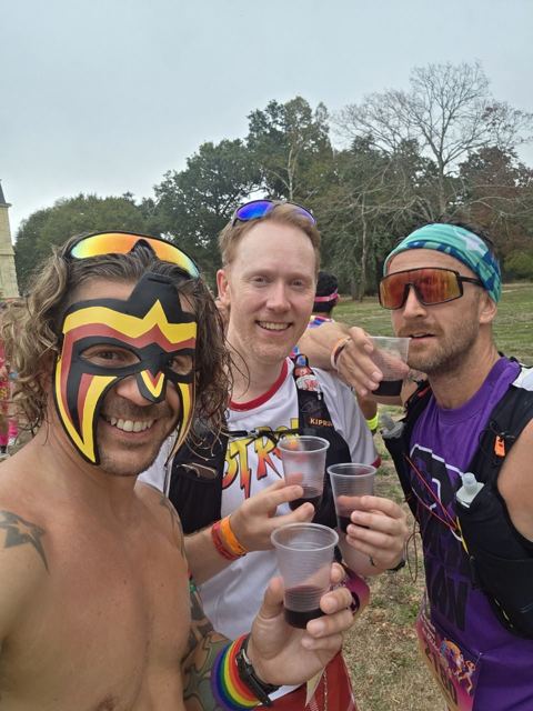 Three runners in costume holding cups of red wine under trees.