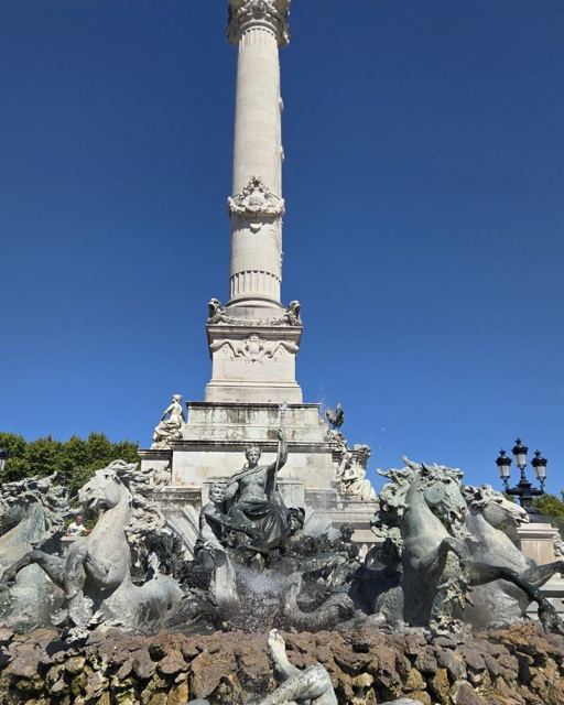 Tall column topped by a statue rising above a fountain of bronze horses under a blue sky.