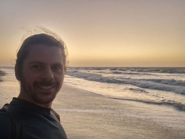 A smiling man taking a selfie on a beach with the sun setting over the sea.