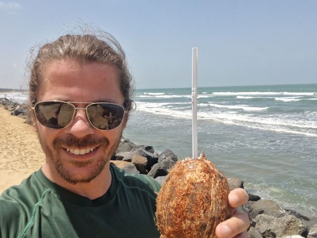 A smiling man in sunglasses holding a coconut with a straw by the sea.
