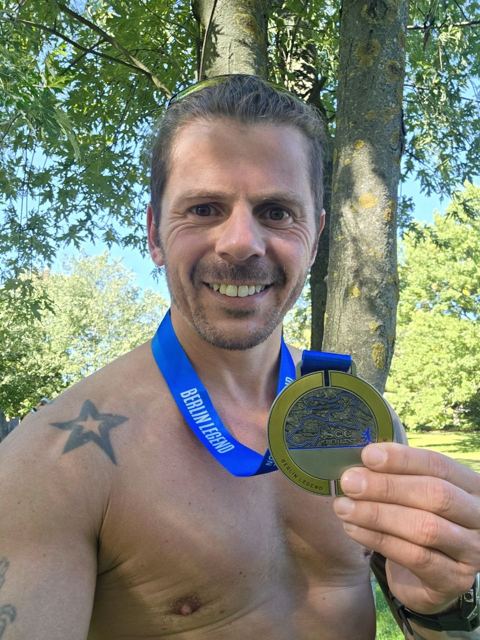 A grinning shirtless man holding up his Berlin Marathon medal.