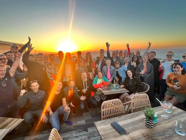 A large group of people cheering at a terrace bar as the sun sets over the sea.
