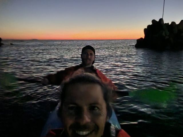 Two people in a kayak on the sea at dusk beside a dark rocky islet.